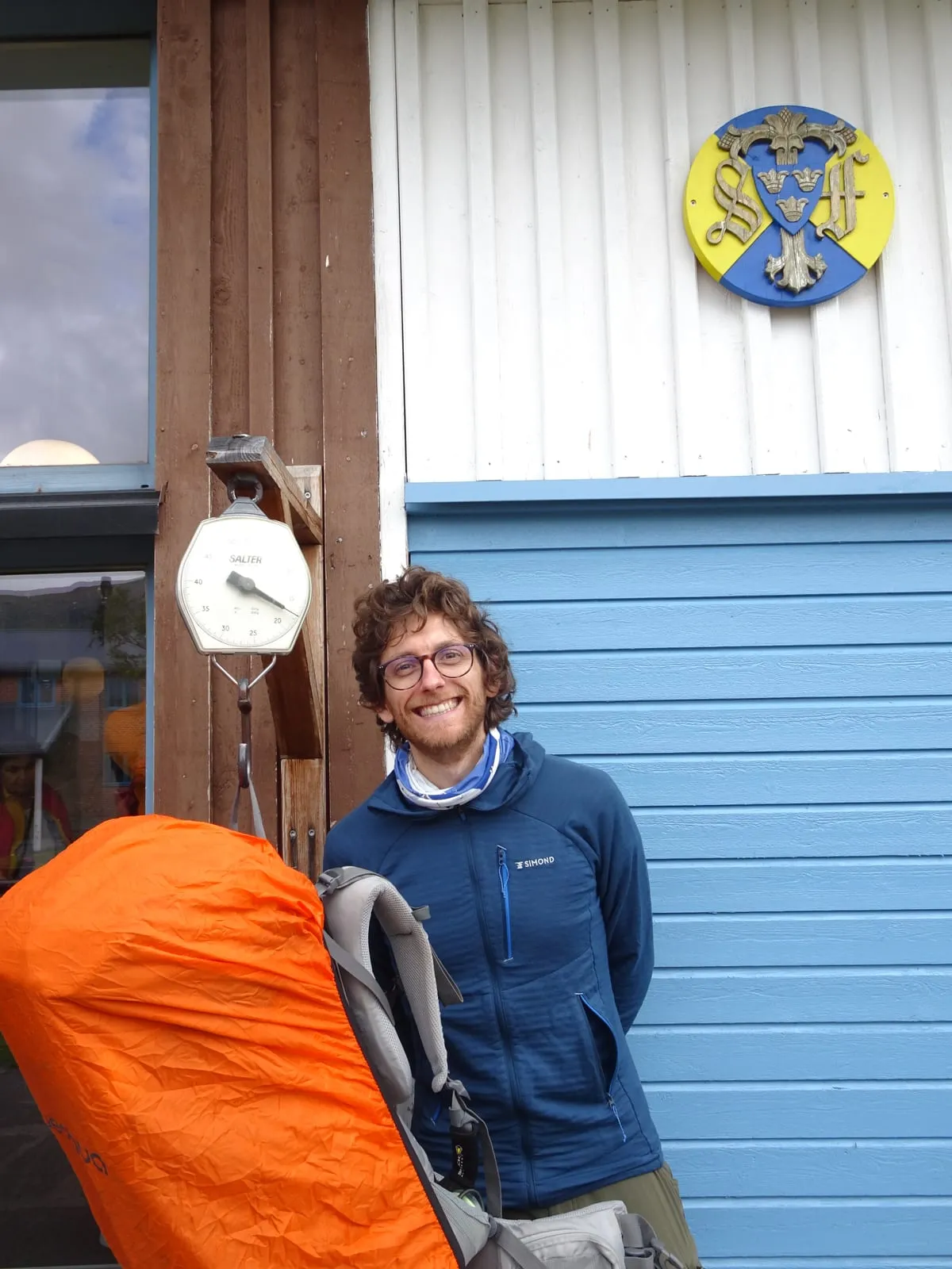 Standing beside a hanging Salter scale at a mountain station, weighing a full pack with an orange rain cover