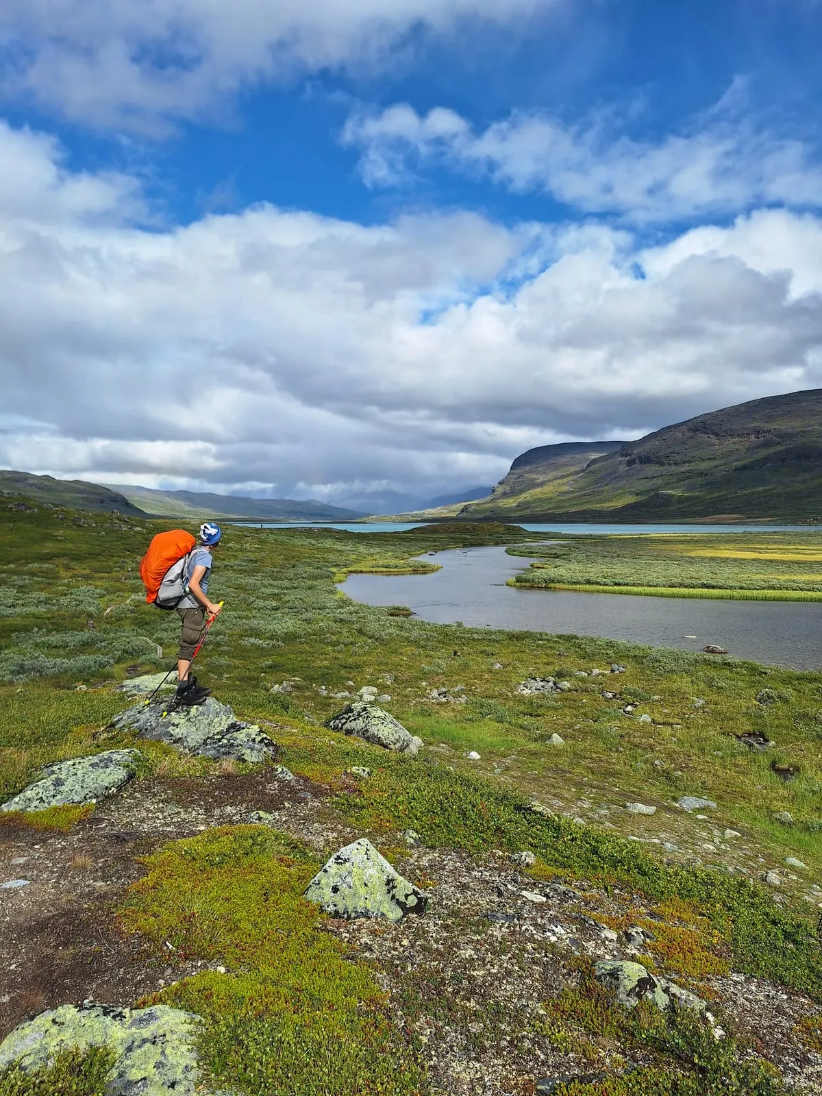 A walker with an orange-covered pack standing on a lichened rock, looking out over a wide valley with a lake and low mountains under a big cloudy sky